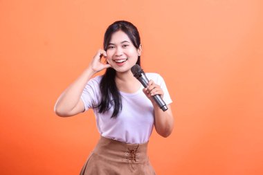 Elinde mikrofon olan ve gülümseyen neşeli genç bir kadın, sahnede turuncu arka planda izole edilmiş, büyük bir coşku ve mutlulukla sahneye çıkıyor gibi görünüyor.