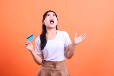 Elinde kredi kartı tutarken üzgün görünen genç bir kadın, kızgın ve hayal kırıklığına uğramış bir yüz ifadesi, turuncu arka planda izole edilmiş.