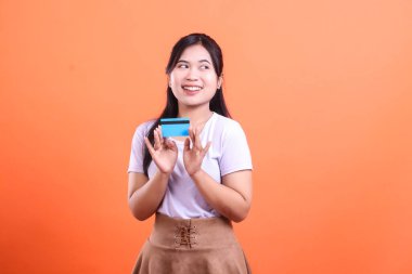 Genç bir kadın elinde mavi bir kredi kartı tutarken gülümsüyor, iki eliyle kartı gösteriyor, turuncu arka planda alışverişe gitmek için heyecanlı.