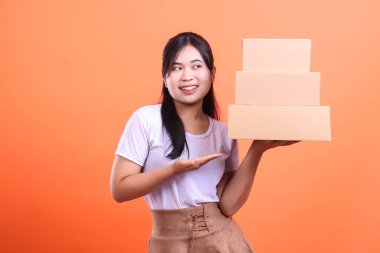 Genç bir kadın karton kutular sunuyor, bir eliyle tutuyor, diğer eliyle işaret ediyor, turuncu arka planda izole edilmiş..