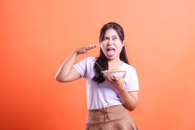 Genç bir Asyalı kadın tiksindirici bir ifade sergiliyor, elinde bir kase yemek, dili açık, turuncu arka planda izole edilmiş bir hoşnutsuzluk veya hoşnutsuzluk hissi veriyor.