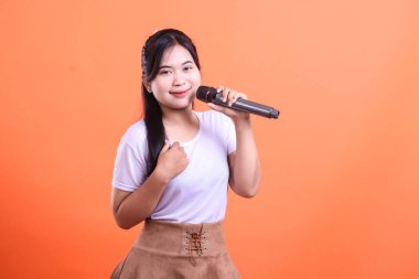 Genç bir Asyalı kadın bir mikrofon tutuyor ve gülümseyerek şarkı söylüyor. Performansından çok emin, turuncu arka planda izole edilmiş.