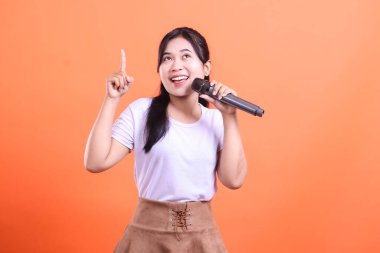 Elinde mikrofon tutan ve parmağını yukarı gösteren gülümseyen genç bir kadın turuncu arka planda mutlu ve neşeli görünüyor..
