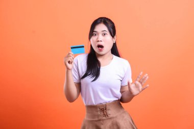 Genç bir kadın kredi kartını elinde tutarken ağzı açık ve elini kaldırmış turuncu arka planda izole bir şekilde şok jesti yaparken son derece şaşırmış görünüyor.
