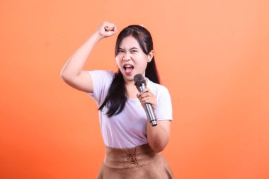 Mikrofon tutan genç bir kadın, tutkuyla şarkı söylüyor, kolları havada, turuncu arka planda izole edilmiş neşeli ve anlamlı bir enerji gösteriyor.