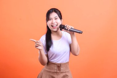 Gülümseyen genç bir kadın mikrofona şarkı söylüyor, solunu mutlu bir ifadeyle işaret ediyor, turuncu arka planda izole edilmiş.