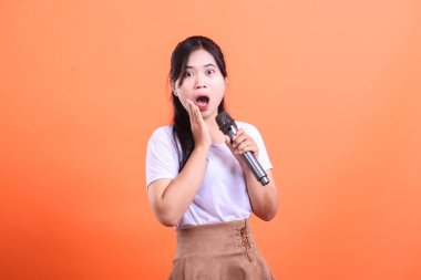 Şok olmuş gibi görünen genç bir kadın, şarkı söylerken elinde bir mikrofon tutuyor, turuncu arka planda izole bir şekilde.