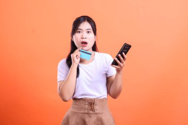 Şaşırmış genç bir kadın kredi kartı ve cep telefonunu elinde tutarken çok şaşırmış görünüyor. turuncu arkaplanda izole