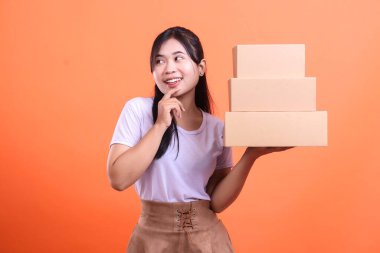 Genç Asyalı bir kadın elinde bir yığın karton kutu tutuyor, düşünceli bir şekilde sağına bakıp gülümsüyor. turuncu arkaplanda izole