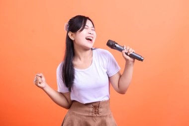 Genç, mutlu bir kadın mikrofon ve enerjik bir ifadeyle şarkı söylüyor. Performansının tadını çıkarıyor. Turuncu arka planda izole bir şekilde.