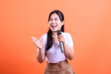 Gülümseyen genç bir kadın mikrofona şarkı söylüyor, bir elini havaya kaldırıyor, turuncu arka planda izole bir şekilde.