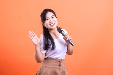 Mikrofonlu genç bir Asyalı kadın büyük bir gülümsemeyle elini sallıyor. Mutlu ve neşeli görünüyor, turuncu arka planda soyutlanmış.