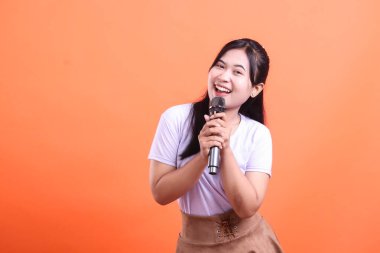 Uzun siyah saçlı neşeli bir Asyalı kadın mikrofonu tutuyor ve şarkı söylerken geniş bir gülümseme sergiliyor. turuncu arkaplanda izole.