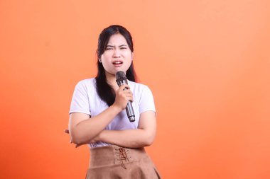 Asyalı bir kadın canlı performans sergilerken yüzünde karışık bir ifade olan bir mikrofon tutuyor. turuncu arkaplanda izole