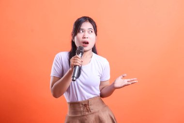 Şaşırmış bir kadın mikrofon tutuyor ve konuşurken jestler yapıyor. Turuncu arka planda izole edilmiş bir tişört ve etek giyiyor..