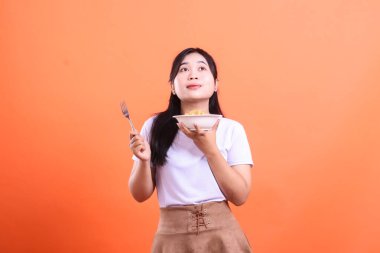 Genç bir kadın elinde bir kase erişte ve çatal tutarken, mutlu ve düşünceli görünüyor, pozitif bir ifadeyle, turuncu arka planda izole edilmiş.