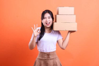 Gülümseyen genç bir kadın, bir elinde kahverengi kutular tutarken diğer elinde turuncu arka planda izole bir şekilde 