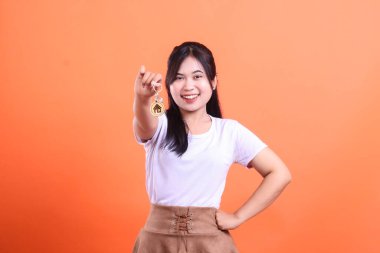 Neşeli genç Asyalı kadın gülümserken evinin anahtarlarını gösteriyor. Turuncu arka planda izole edilmiş emlak, satın alma veya yeni bir daire kiralama kavramı.