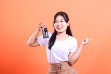 Gülümseyen ve elinde bir araba anahtarı tutan neşeli bir kadın yön için bir tarafı işaret ediyor. turuncu arkaplanda izole.