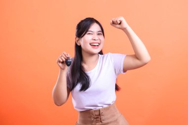 Neşeli, genç bir kadın sevinçli bir ifadeyle araba anahtarlarını tutuyor ve başarısını gösteren yumruğunu kaldırıyor. turuncu arkaplanda izole