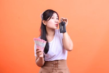 Heyecanlı genç bayanın elinde nakit para ve araba anahtarı var. Bu da mali başarı ve yeni bir satın alma olduğunu gösteriyor. turuncu arkaplanda izole.