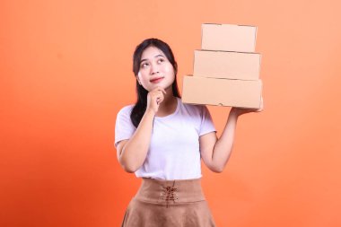 Genç bir kadın elinde üç karton kutu tutarken yukarı bakıyor, internetten siparişleri ya da teslimatları hakkında kafa yoruyor, turuncu arka planda izole edilmiş..