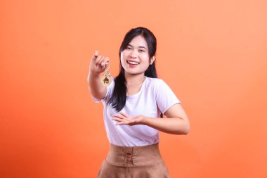 Neşeli genç bir kadın ev sahipliğini ve yeni başlangıçları simgeleyen bir ev anahtarını gururla sunar. Turuncu arka planda izole edilmiş yeni evinin sembolünü göstererek geniş bir gülümseme sergiliyor..