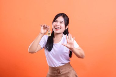 Neşeli bir Asyalı kadın, önemli bir başarı ve gelecekteki refahı simgeleyen yeni evinin anahtarlarını gururla sergiliyor. turuncu arkaplanda izole.