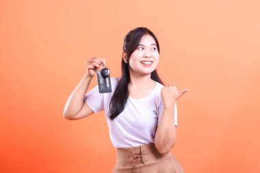 Mutlu genç Asyalı kadın arabanın anahtarlarını tutuyor ve bir gülümsemeyle yan tarafı işaret ediyor. turuncu arkaplanda izole.