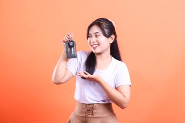 Gülümseyen genç bayan yeni araba anahtarlarını el hareketiyle sunar. Turuncu arka planda izole edilmiş neşeli ve kendinden emin bir araç sahibi anı..