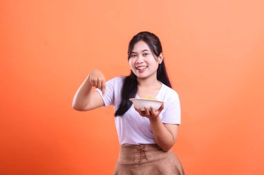 Genç ve güzel bir kadın bir kase erişteyi sunarken gülümsüyor, sanki size gösteriyormuş gibi, turuncu arka planda izole bir şekilde.