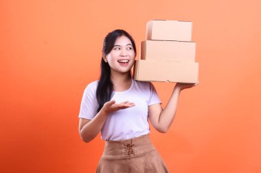 Elinde bir yığın karton kutu tutan neşeli genç bir kadın. Turuncu arka planda teslimat, dağıtım ya da satın almaya hazır.