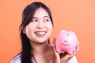 Elinde pembe bir kumbarayla gülümseyen Asyalı bir kadın umut dolu bir ifadeyle yan tarafa bakıyor. Turuncu arka planda mali tasarruf ve planlama teşviki.