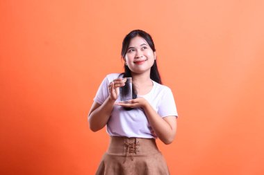 Bir bardak su tutan, sıradan giysili çekici bir kadın. Düşünceli bir şekilde yukarı bakıyor. turuncu arkaplanda izole.