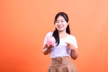 Elinde tuttuğu pembe domuz kumbarasını işaret eden gülümseyen genç Asyalı kadın, turuncu arka planda izole edilmiş mali sorumluluğun sembolü.