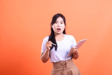 Şaşırtıcı bir ifade, konsept teknolojisi ve dijital iletişimi olan bir tablet kullanan genç bir kadın, turuncu arka planda izole edilmiş..