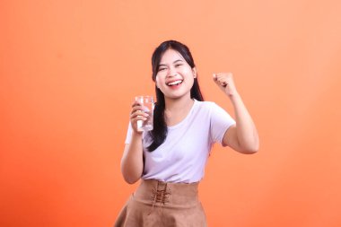 Neşeli genç bir kadın bir bardak temiz su tutuyor ve stüdyoda gülümsüyor. Sağlığa ve vücuda iyi gelir. turuncu arkaplanda izole.