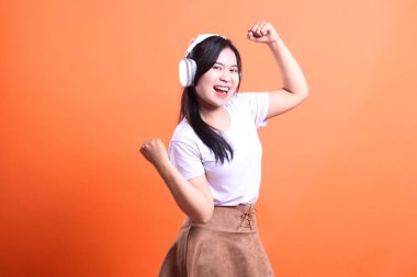 Beyaz kulaklık, beyaz tişört ve kahverengi etek giyen genç bir kadın turuncu arka planda izole bir şekilde kollarını kaldırıyor..