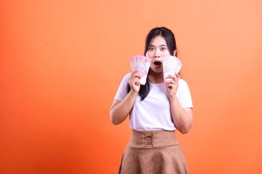 Siyah saçlı genç bir kadın elinde iki para fanatiği tutuyor ve ağzı açık bekliyor. O, beyaz bir tişört ve kahverengi bir etek giyiyor ve parlak turuncu bir arka planın karşısında duruyor..