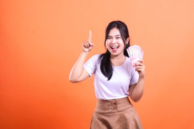 Siyah saçlı, gülümseyen bir kadın, Endonezya Rupiah banknotlarını tutarken turuncu bir arka plana doğru işaret ediyor..