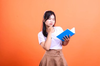 Koyu saçlı bir kadın turuncu arka planda dururken mavi bir kitap tutuyor. Beyaz bir tişört ve kahverengi bir etek giyiyor. turuncu arkaplanda izole