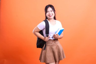 Sırt çantalı genç bir kadın elinde mavi bir kitapla turuncu arka plandaki kameraya doğru gülümsüyor.