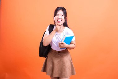 Sırt çantası ve mavi kitabı olan gülümseyen bir öğrenci başka tarafa bakıyor. Günlük kıyafetler giyiyor ve turuncu arka planda izole edilmiş turuncu bir arka planın önünde duruyor.