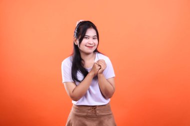 Turuncu arka planda bir kadın ayakta ve gülümsüyor. Koyu renk saçları var, beyaz gömlek ve kahverengi bir etek giyiyor..