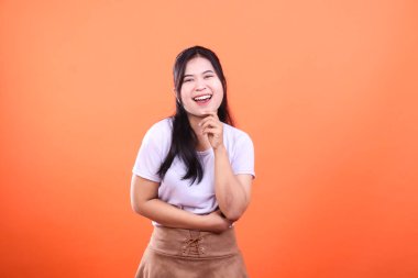 Siyah saçlı ve beyaz tişörtlü, turuncu arka planda parlak bir şekilde gülümseyen genç bir kadının portresi. Eli çenesinde, neşeli ve olumlu bir sahne yaratıyor..