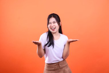 Genç bir kadın, elleri açık gülümseyip göz kırpıyor. İki avucunda da bir şey var. O, turuncu bir arka planda izole edilmiş beyaz gömlek ve kahverengi bir etek giyiyor..