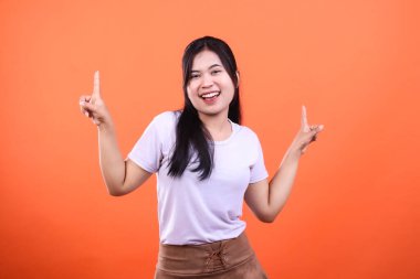 Koyu renk saçlı ve açık renk gömlekli bir kadın iki eliyle yukarı bakıyor, mutlu bir şekilde gülümsüyor. Kahverengi bir etek giyiyor ve turuncu bir arka planda izole edilmiş..