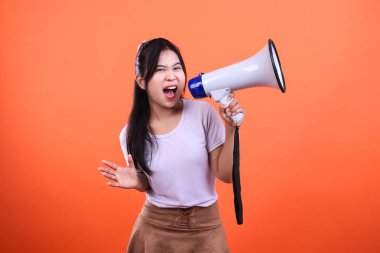 Bir kadın, eli açık, turuncu bir arka planda izole bir şekilde kameraya bakarken beyaz ve mavi bir megafona bağırıyor..