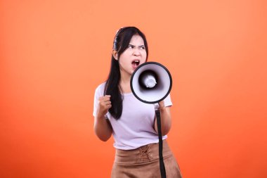 At kuyruklu siyah saçlı bir kadın elinde megafonla bağırıyor. O, beyaz bir gömlek ve kahverengi bir etek giyiyor. Yüzünde kararlı bir ifade var. Arka plan turuncu. Turuncu bir arka planda izole edilmiş..