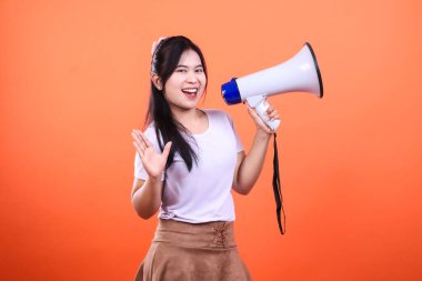 Siyah saçlı bir kadın beyaz ve mavi bir megafon tutuyor ve elini sallıyor, gülümsüyor. Beyaz bir gömlek ve kahverengi bir etek giyiyor, turuncu bir arka planda izole edilmiş..